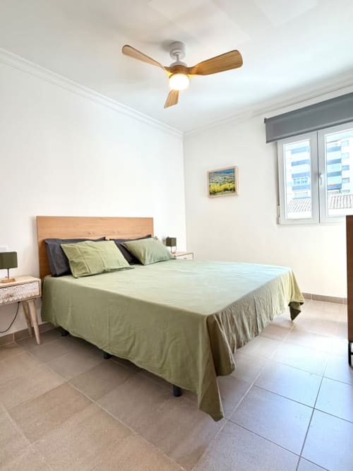 Dormitorio luminoso en casa rural Casa Mar Blau, Valencia, con cama de madera, ropa de cama verde, ventilador de techo y vistas al mar