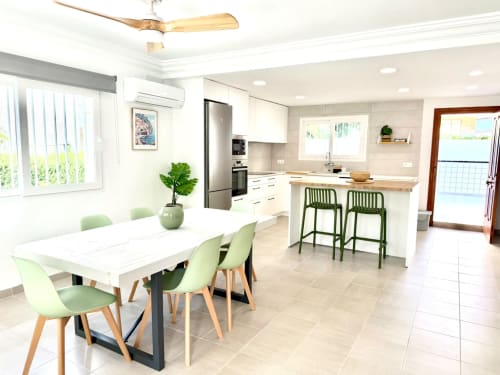 Cocina comedor de planta abierta en casa de playa Casa Mar Blau, Valencia, con muebles blancos, taburetes verdes y vistas al mar