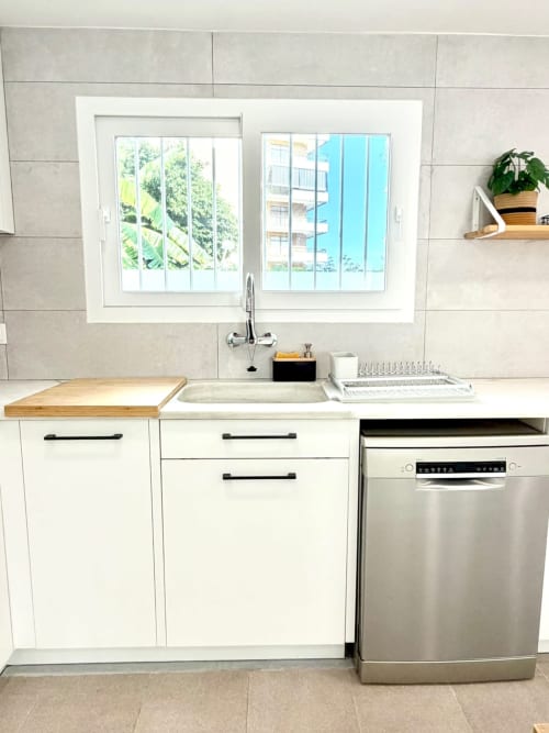 Cocina moderna con muebles blancos, lavavajillas de acero inoxidable y ventana con vistas al mar en Casa Mar Blau