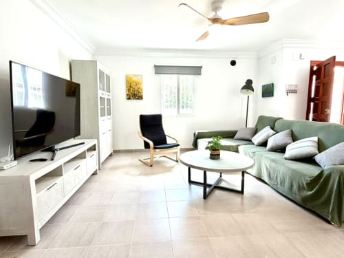 Salón luminoso y moderno en casa de campo Casa Mar Blau con sofá verde, televisor, ventilador de techo de madera y mobiliario blanco en Valencia