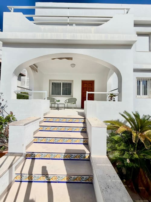Entrada de acogedora casa mediterranea blanca con escalones decorativos de azulejos azules y amarillos, pórtico abovedado y palmeras