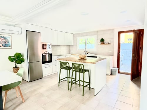 Cocina moderna blanca con isla, taburetes verdes, electrodomésticos de acero inoxidable y vistas al mar en Casa Mar Blau,