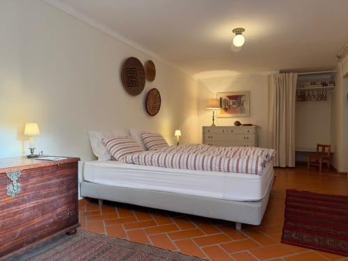 Quarto luminoso na Quinta da Mata de Baixo com cama queen, roupas de cama listradas em vermelho, baú de madeira e piso de azulejo cerâmico