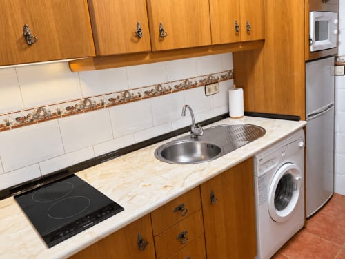 Cocina con armarios de madera, salpicadero de azulejos blancos, encimera de mármol, placa de inducción y lavadora