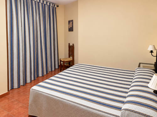 Luminosa habitación del hotel La Posada en Oropesa con ropa de cama rayada, cortinas azules, muebles de madera y piso de terracota