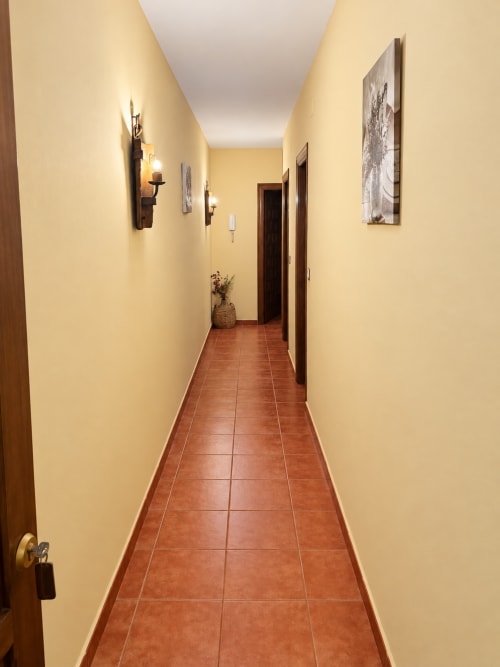 Pasillo largo y estrecho con paredes color crema, piso de baldosas terracota, puertas de madera y apliques de pared en Apartamentos La Posada