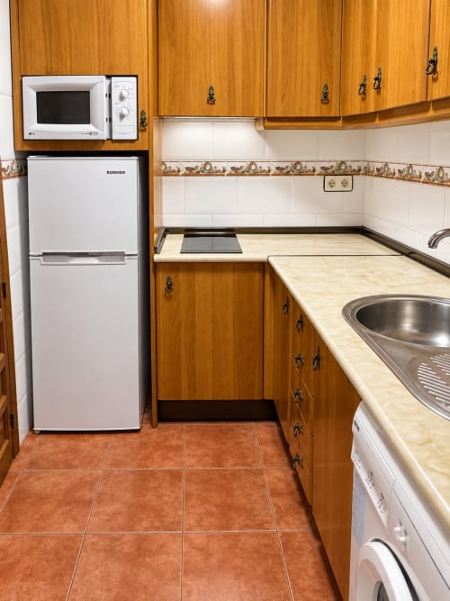 Cocina compacta en estudio de La Posada, Oropesa con armarios de madera, refrigerador blanco y piso de barro