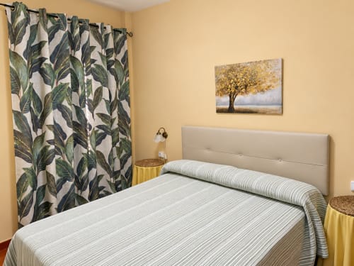 Dormitorio de los apartamentos La Posada en Oropesa con ropa de cama a rayas, cortinas botánicas y obra de arte de árbol dorado