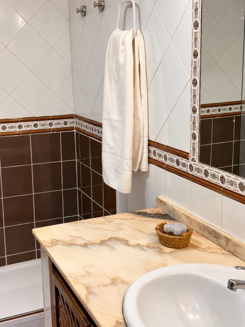 Baño en Apartamentos La Posada, Oropesa Toledo - encimera de mármol, paredes con azulejos marrones, toallas blancas en el toallero