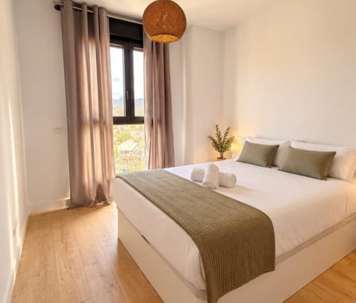 Luminoso dormitorio moderno en Sinese Design Homes Málaga con vistas a la montaña, suelos de madera, cama blanca, almohadas verdes y tonos beige