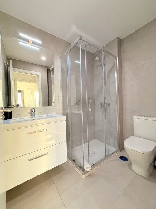 Baño moderno en apartamento de Sinese Design Homes en Málaga con vanidorio blanco, ducha con mampara de vidrio, inodoro y piso climatizado