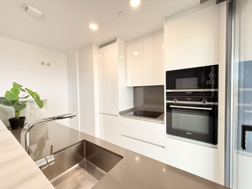 Moderna cocina blanca con fregadero de acero inoxidable, electrodomésticos negros y planta en maceta en apartamento de Martiricos, Málaga