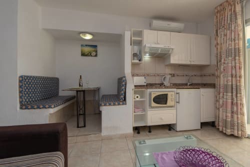 Modern studio kitchenette with white cabinets, microwave, and dining area with blue patterned seating in Costa Adeje