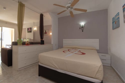 Modern studio bedroom in Costa Adeje with white furniture, purple accent wall, ceiling fan, and pool views from balcony