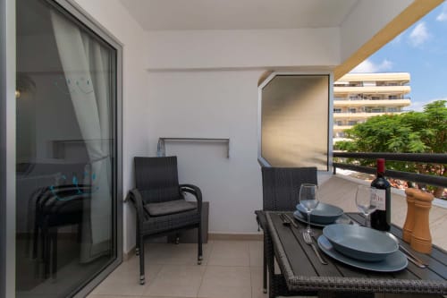 Sunny balcony in Costa Adeje with dining table, wicker chairs, wine bottle, and apartment building view