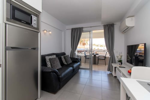 Modern studio apartment in Costa Adeje with black sofa, kitchenette, TV, and balcony overlooking pool and sea