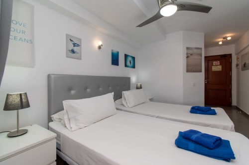 Modern studio bedroom in Costa Adeje with two beds, gray headboard, blue towels, ocean-themed wall art, and ceiling fan
