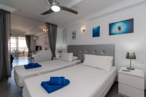 Modern studio apartment in Costa Adeje with twin beds, gray headboard, ocean-themed artwork, ceiling fan, balcony access, and