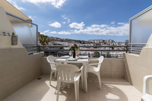 Sunny balcony with white furniture overlooking Costa Adeje, Tenerife. Dining table, chairs, and sea view of hillside town.