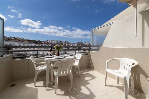 Sunny balcony with white furniture overlooking Costa Adeje, Tenerife. Dining table, chairs, sea views, and coastal cityscape