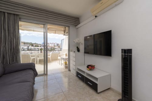 Modern studio apartment in Costa Adeje with flat-screen TV, white furniture, balcony with sea view, and tiled flooring.