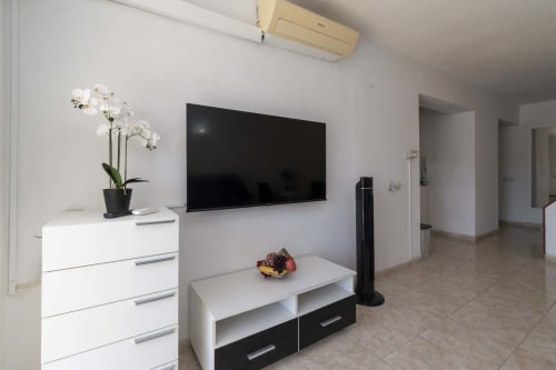 Modern studio apartment living room with flat-screen TV, white furniture, orchid plant, and air conditioning unit in Costa