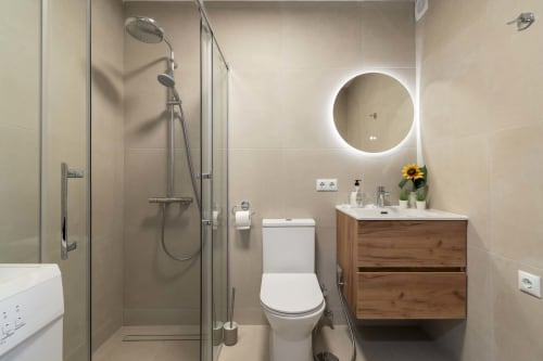 Modern bathroom in Costa Adeje studio with illuminated circular mirror, wooden vanity, walk-in shower, and white toilet.