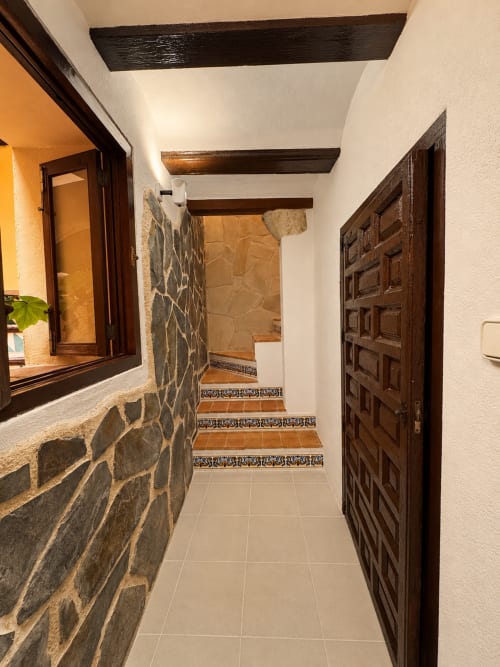 Corredor de piedra rústica con vigas de madera oscura, puerta ornamentada y escaleras con azulejos decorativos en Casa Rural Garden