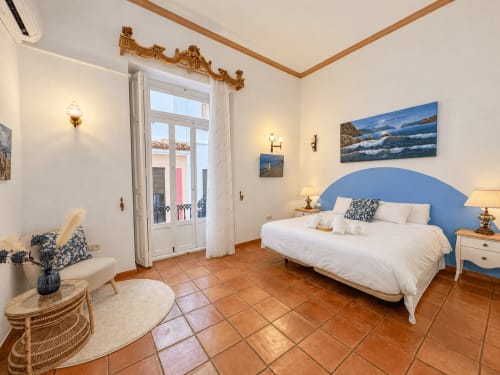 Amplio dormitorio con cabecero azul, suelo de barro, puertas de balcón y arte costero en Casa Rural Garden Suites