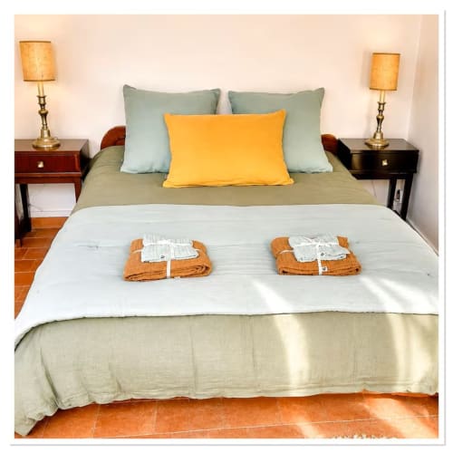 Chambre accueillante au cottage Les Maraîchades avec literie sauge, coussin jaune, lampes jumelles et carrelage terre cuite.