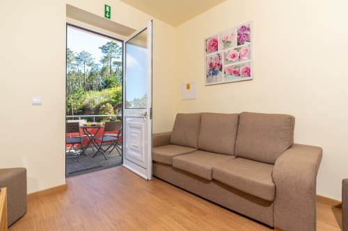 Bright living room with beige walls, tan sofa, wooden floors, and open glass door leading to terrace with mountain views at