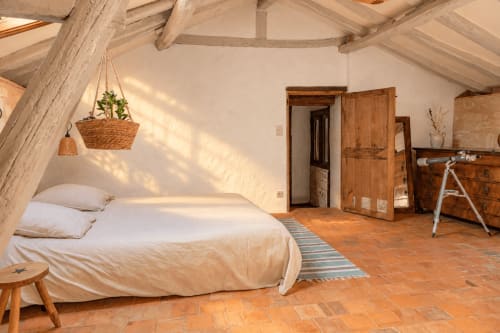 Chambre rustique au gîte Le Clos Divin avec poutres apparentes, lit blanc, carrelage en terre cuite, portes en bois et plantes suspendues