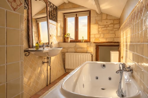 Salle de bain élégante au gîte Le Clos Divin avec baignoire îlot, lavabo sur piédestal et murs en pierre chaleureuse inondés de lumière naturelle