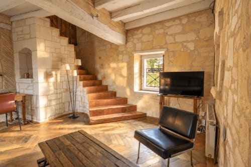 Intérieur rustique de la maison en pierre du Clos Divin avec poutres en bois, escalier en terre cuite, télévision et vue sur le jardin à Doué-la-Fontaine