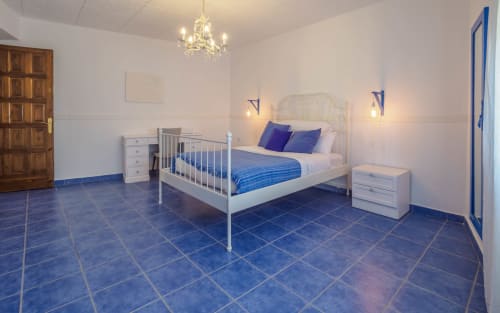 Amplio dormitorio de villa en Masias Can Trabal con suelo de azulejos azules, cama blanca, lámpara de araña y vistas al jardín en Olèrdola