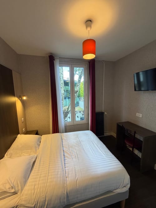Chambre lumineuse à l'Hôtel le Dellys à Colombes avec lit blanc, luminaire rouge, portes-fenêtres et télévision