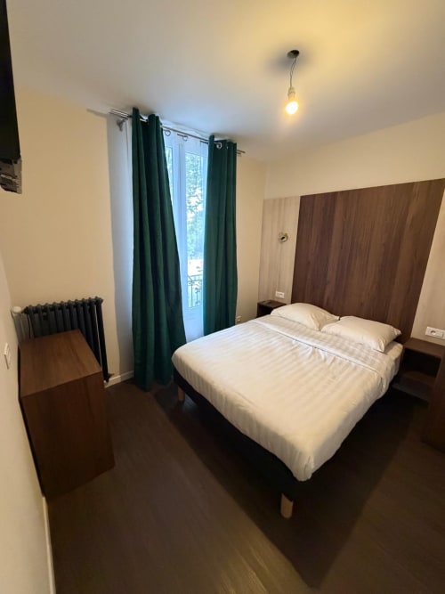 Chambre moderne de l'Hôtel le Dellys à Colombes avec lit queen-size, tête de lit en bois foncé, rideaux verts et luminaire suspendu