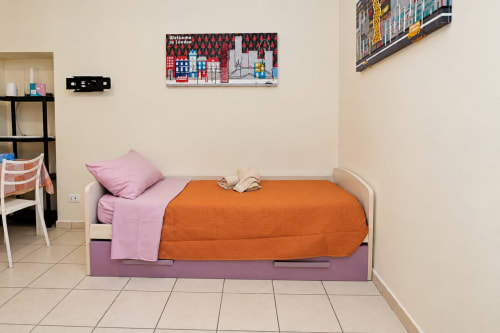 Camera luminosa con letto singolo a Sidra Apartments Bari, coperta trapuntata arancione, biancheria rosa e opere d'arte con vista città su beige