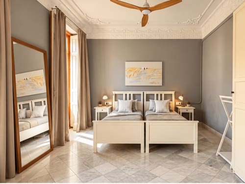 Lujoso dormitorio de apartamento en Valencia con camas gemelas, muebles color crema, paredes grises, elegante ventilador de techo y cortinas con vista a la ciudad