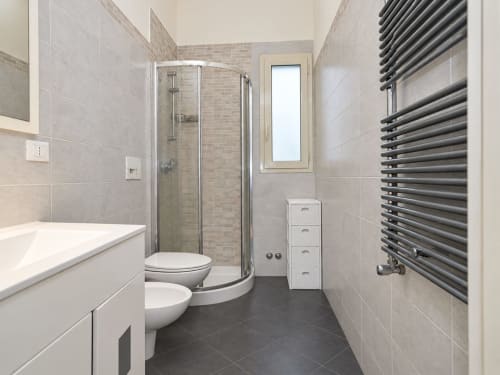 Bagno moderno degli Sidra Apartments a Bari con doccia curva, wc, bidet, scaldasalviette e pavimento in piastrelle scure.