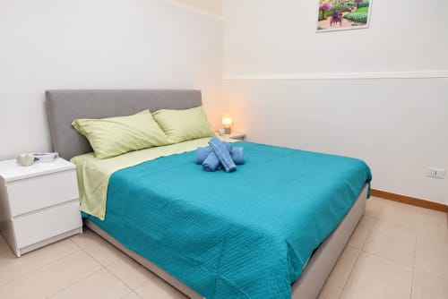 Camera luminosa negli Sidra Apartments di Bari con coperta turchese, cuscini verde lime, testata grigia e comodino bianco