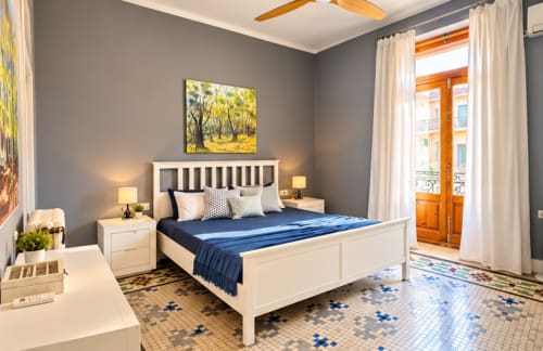 Dormitorio de lujo en apartamento Stay Gest Valencia con cama blanca, acentos azules, ventilador de techo de madera y vistas a la ciudad desde balcón