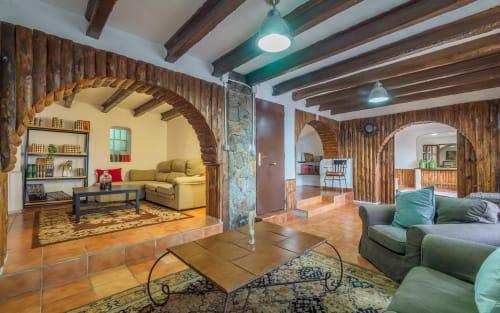 Amplia sala de estar de la villa Masías Can Trabal con alcoba de arco de ladrillo, vigas de madera, asientos cómodos y montaña