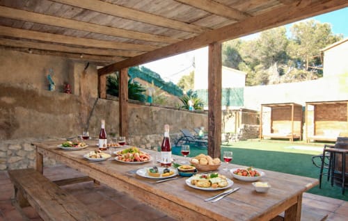 Comedor al aire libre en villa Masías Can Trabal con mesa de madera rústica, platos mediterráneos, botellas de vino y montaña