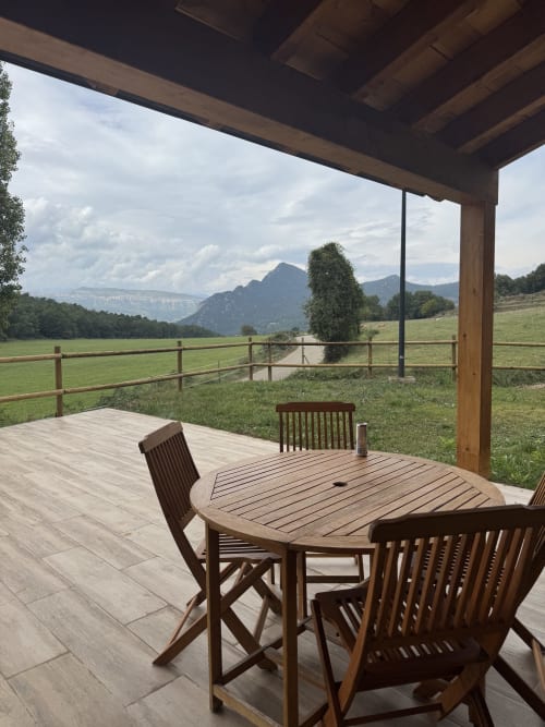 Patio cubierto con mesa de comedor de madera y sillas con vistas a los campos verdes y montañas de la casita Cal Boix, Berga