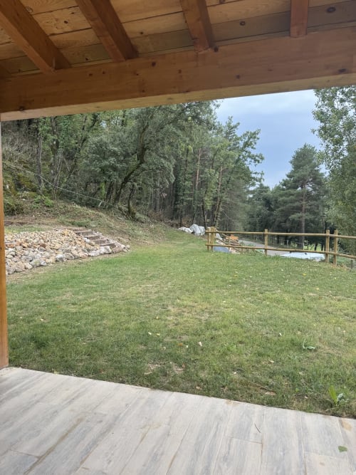 Terraza de madera con vistas al jardín paisajístico, césped, sendero de piedra, barandillas de madera y montañas boscosas en Cal Boix