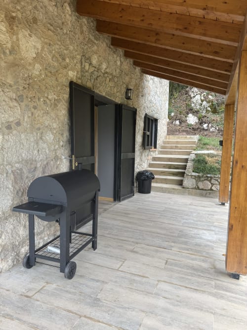 Patio cubierto de la casita Cal Boix en Berga con parrilla BBQ negra, muros de piedra y vigas de madera con vistas a la montaña
