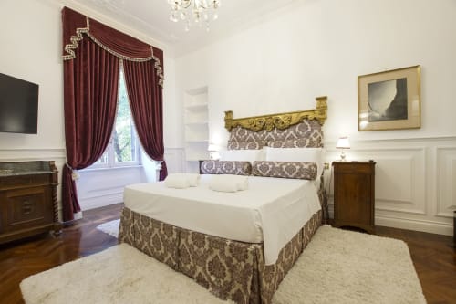 Elegante camera da letto della Suite Magna Mater a Roma con arredi ornati, tende bordeaux e arredamento classico