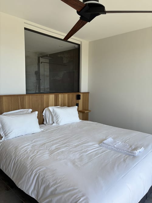 Chambre moderne aux Villas d'A Storia à Pinarellu avec tête de lit en bois, literie blanche, ventilateur de plafond et salle de bains attenante