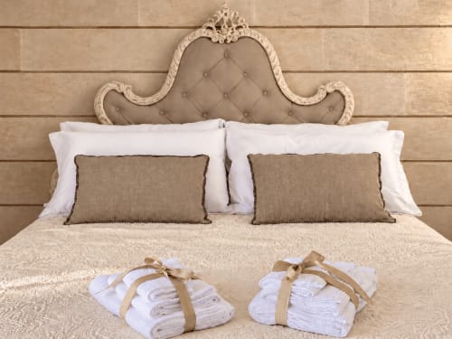 Camera elegante a HABITARE Lecce con testiera ornata, cuscini bianchi, cuscinetti beige e asciugamani arrotolati legati con nastro dorato